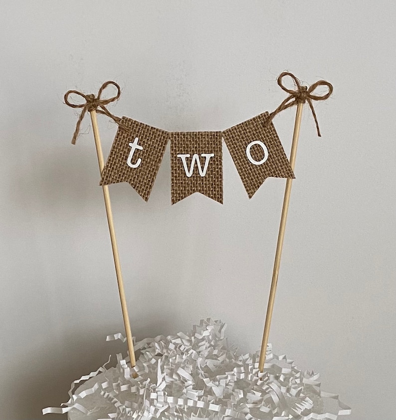 Puede incluir: Adorno para tarta con una pancarta de arpillera con la palabra "TWO" en letras blancas. La pancarta est&aacute; suspendida entre dos palitos de madera, cada uno adornado con un lazo de cuerda. El adorno est&aacute; sobre virutas de papel blancas.