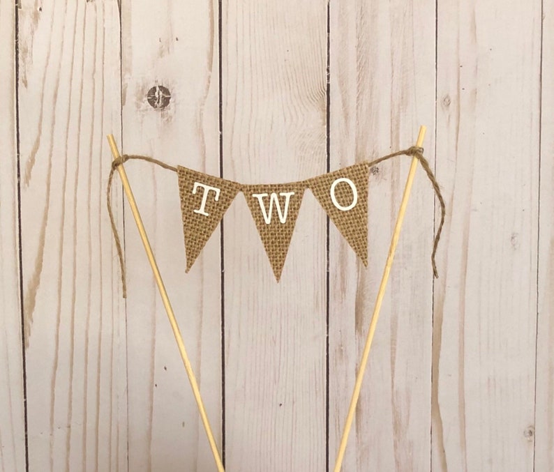 Puede incluir: Un adorno para tarta con forma de bander&iacute;n r&uacute;stico de arpillera con la palabra "TWO" en letras blancas. El bander&iacute;n est&aacute; ensartado en hilo y sujeto a dos palillos de madera, ideal para una celebraci&oacute;n de segundo cumplea&ntilde;os.