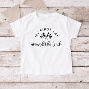 May include: White t-shirt with black text that reads "My First Lap Around the Track" with a checkered flag graphic.
