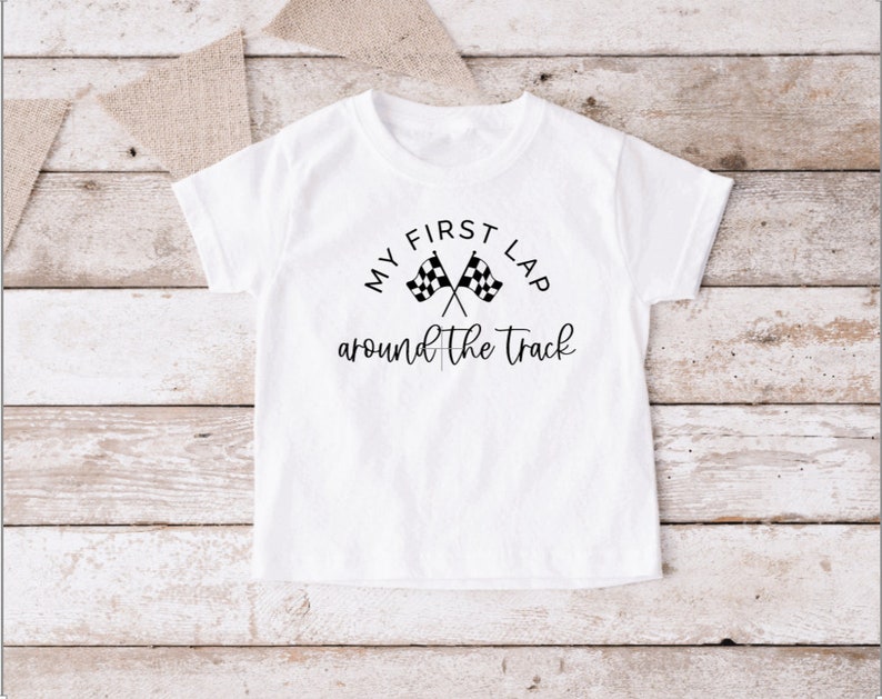 May include: White t-shirt with black text that reads "My First Lap Around the Track" with a checkered flag graphic.
