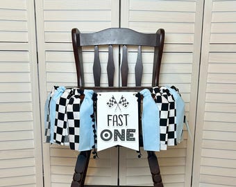 May include: A high chair banner with a racing theme. The banner features a black and white checkered pattern, light blue fabric, and a white pennant that reads "FAST ONE" with checkered flags. Black pom-poms trim the banner.