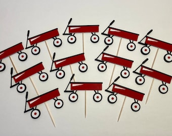 May include: Twelve red and black wagon cupcake toppers with white wheels. The toppers are on wooden skewers.