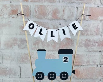 May include: A cake topper featuring a light blue train engine with three black wheels and the number 2 on its side. Above the train, a white bunting banner hangs from two wooden sticks, displaying the name OLLIE in bold black capital letters.