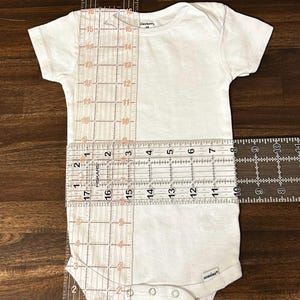 May include: White onesie with a round neckline and snap closures. The onesie is measured with rulers, showing a length of 17 inches and a width of 10 inches.