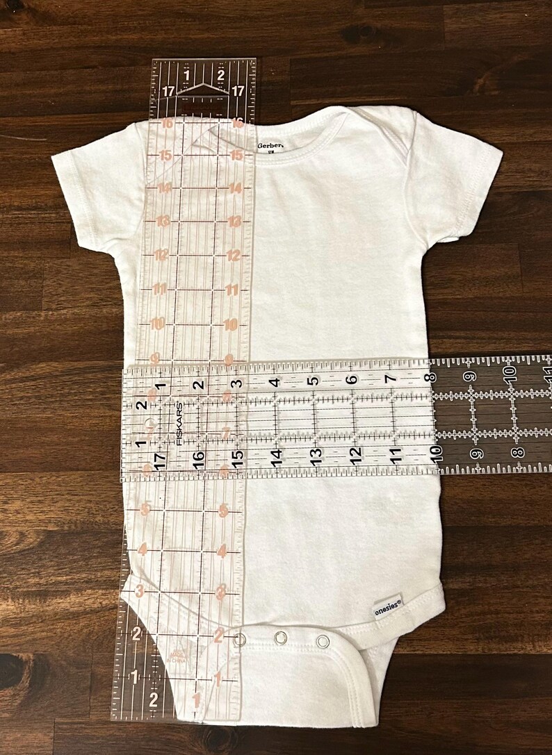 May include: White onesie with a round neckline and snap closures. The onesie is measured with rulers, showing a length of 17 inches and a width of 10 inches.