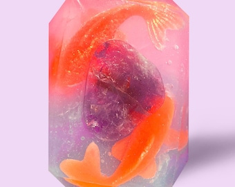 May include: A clear, pink, and purple soap bar with two orange goldfish inside. The soap bar is shaped like a gemstone.