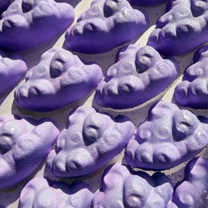 May include: A collection of purple bath bombs shaped like clouds. The bath bombs have a sparkly, iridescent finish.