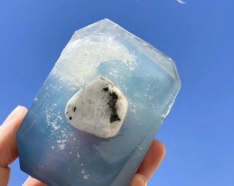 May include: A clear blue resin block with a white and black moonstone embedded inside. The block is being held in a hand against a blue sky background.