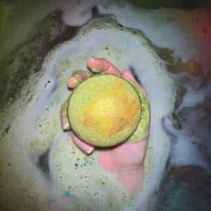 May include: A round bath bomb with a yellow and green exterior and a gold center is held in a hand, surrounded by bubbly, green-tinted water. The bath bomb appears to be dissolving, creating a foamy, colorful bath experience.