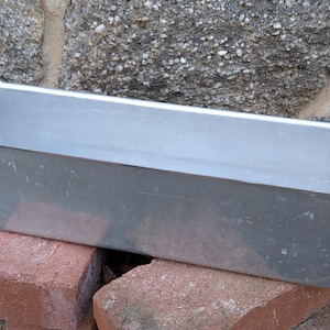 May include: A rectangular, silver-colored metal container with curved handles on each end. The container is placed on top of two red bricks, with a stone wall in the background. The container appears to be a planter or storage bin.