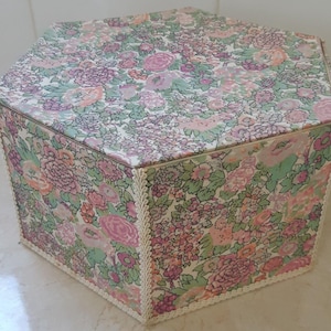 Peut inclure: Une boîte hexagonale à motif floral avec une bordure en dentelle blanche. La boîte est en carton et présente un motif floral sur le dessus et les côtés.