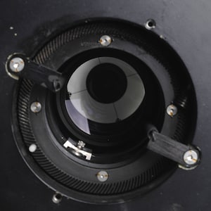 Puede incluir: Vista de cerca de un objetivo de c&aacute;mara negro con una abertura circular y un blanco en el interior. El objetivo tiene un anillo de metal negro con un patr&oacute;n de engranaje y varios tornillos peque&ntilde;os.
