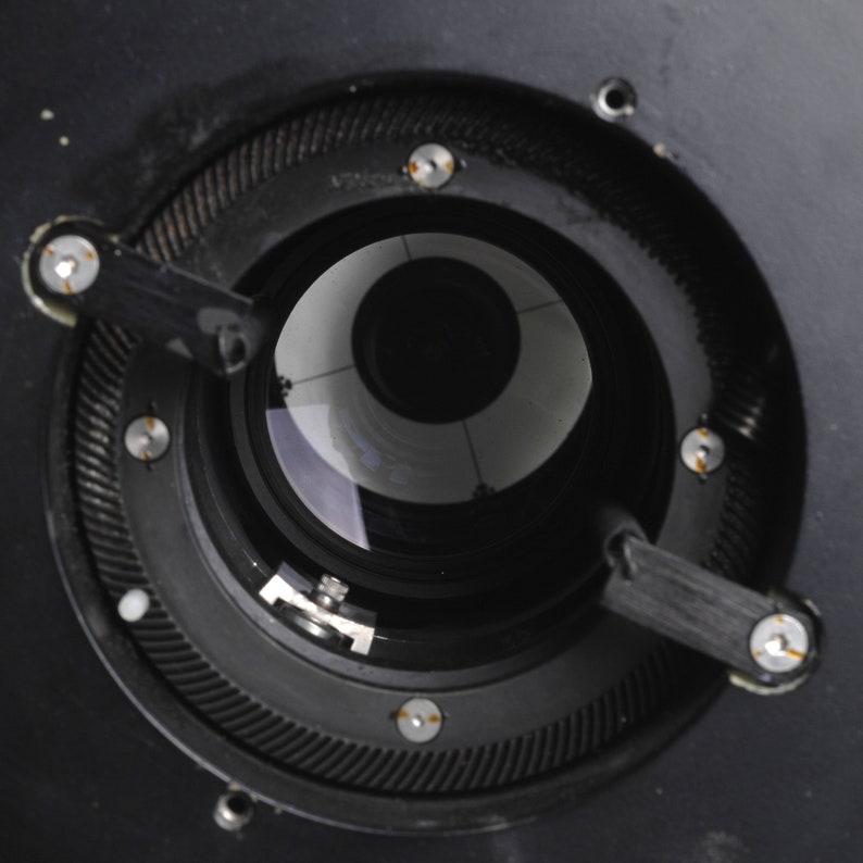 Puede incluir: Vista de cerca de un objetivo de c&aacute;mara negro con una abertura circular y un blanco en el interior. El objetivo tiene un anillo de metal negro con un patr&oacute;n de engranaje y varios tornillos peque&ntilde;os.