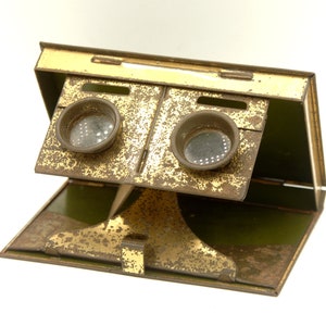 Può includere: Un visore vintage color ottone, portatile, con due lenti rotonde. Il visore è in metallo e ha un aspetto usurato e invecchiato. Il visore è aperto e l'interno è verde.