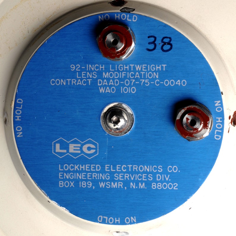 Puede incluir: Una placa met&aacute;lica circular azul con el texto "NO HOLD" impreso en la parte superior e inferior. La placa tiene el texto "92-INCH LIGHTWEIGHT LENS MODIFICATION CONTRACT DAAD-07-75-C-0040 WAO IOIO" impreso en el centro. La placa tambi&eacute;n tiene el texto "LEC LOCKHEED ELECTRONICS CO. ENGINEERING SERVICES DIV. BOX 189, WSMR, N.M. 88002" impreso en el centro. La placa tiene el texto "NO HOLD" impreso en el lado derecho y el texto "OTOH ON" impreso en la parte inferior. La placa tiene el n&uacute;mero "38" impreso en la parte superior derecha.