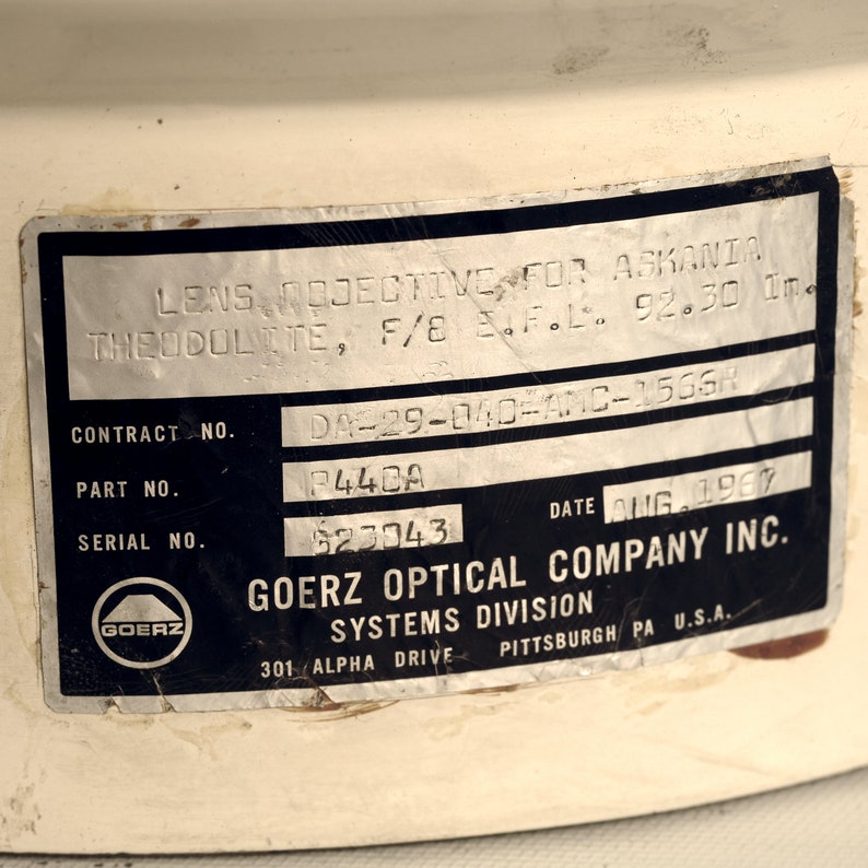 Puede incluir: Una etiqueta en blanco y negro con el texto "GOERZ OPTICAL COMPANY INC. SYSTEMS DIVISION 301 ALPHA DRIVE PITTSBURGH PA U.S.A." y el texto "LENS OBJECTIVE FOR ASKANIA THEODOLITE, F/8 E.F.L. 92.30 In DA-29-040-AMC-15664 CONTRACT NO. P442A PART NO. 623043 SERIAL NO. DATE AUG 1960". La etiqueta tiene un logotipo en blanco y negro de un c&iacute;rculo con la palabra "GOERZ" dentro.