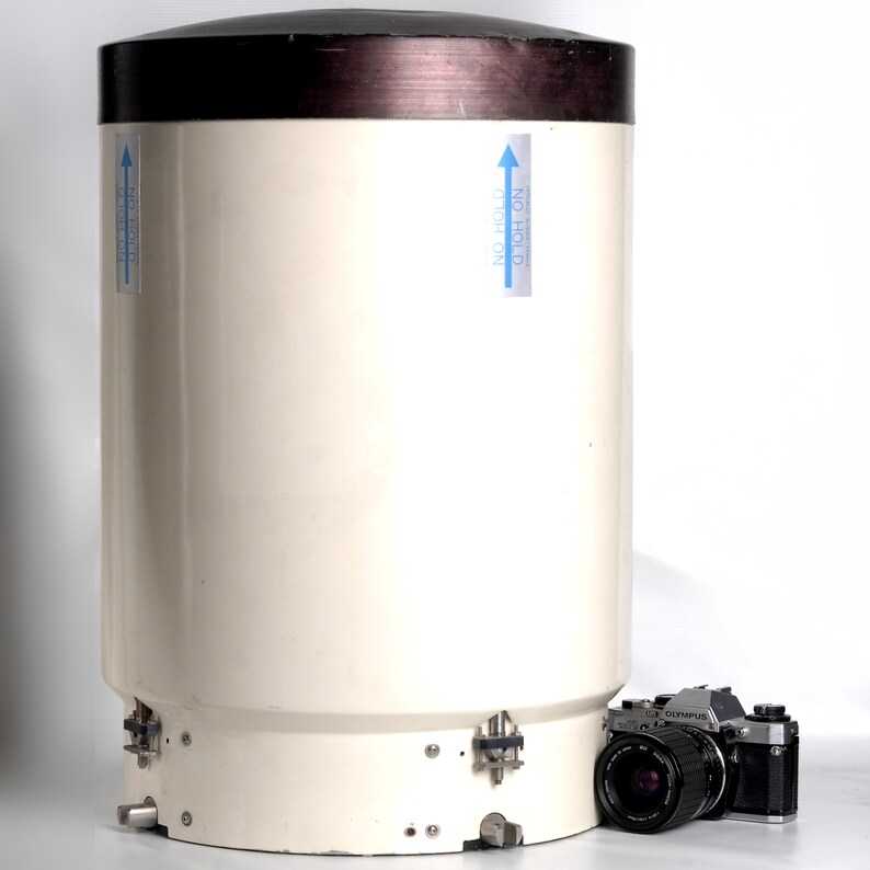 Puede incluir: Una estaci&oacute;n meteorol&oacute;gica cil&iacute;ndrica blanca con una parte superior marr&oacute;n y dos pegatinas azules que dicen "NO HOLD" con una flecha apuntando hacia arriba. Una c&aacute;mara negra y plateada est&aacute; en primer plano.