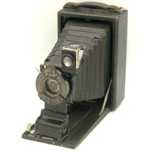 May include: A vintage black folding camera with a bellows system and a lens with a circular aperture. The camera has a leather handle and a metal plate with markings.