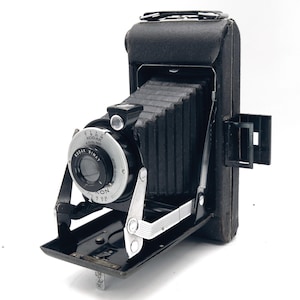 May include: A vintage Kodak folding camera with a black leather-covered body and a black accordion-style bellows. The front features a silver circular lens assembly labeled Kodak Bimat with various aperture and focus settings clearly visible.