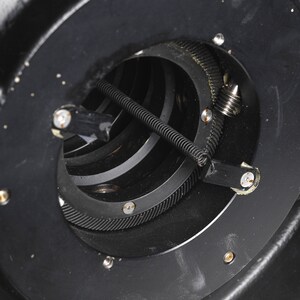 Puede incluir: Primer plano de un sistema de engranajes de metal negro con un resorte y una peque&ntilde;a varilla de metal. Los engranajes son visibles a trav&eacute;s de una abertura circular en el metal.