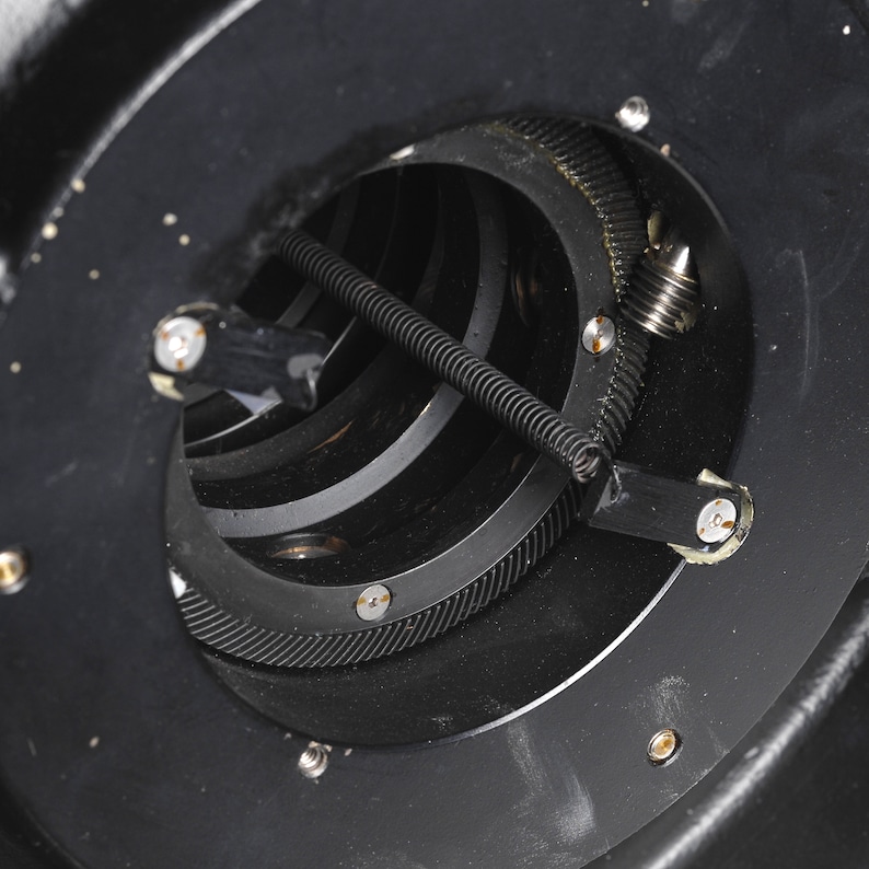 Puede incluir: Primer plano de un sistema de engranajes de metal negro con un resorte y una peque&ntilde;a varilla de metal. Los engranajes son visibles a trav&eacute;s de una abertura circular en el metal.