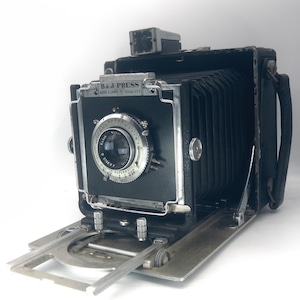 May include: A black B&J Press camera with a folding bellows design, featuring a silver Kodak Supermatic lens assembly on the front panel. The camera includes a top-mounted viewfinder, a metal base plate, and a side handle, with clear text reading B&J PRESS and BURKE & JAMES INC. CHICAGO U.S.A. on the front.