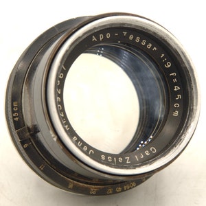 Op de afbeelding: Een zwart en zilveren camera lens met de tekst "Carl Zeiss Jena" en "APO-Tessar 1:9 f=45cm" gedrukt op de zijkant. De lens heeft een scherpstelring met markeringen in centimeters.