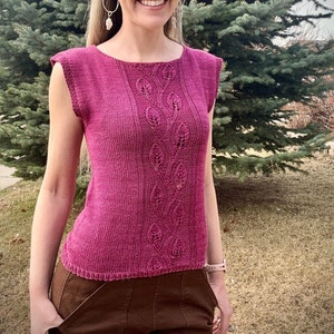 May include: A woman wearing a pink knitted sleeveless top with a leaf pattern and brown pants.