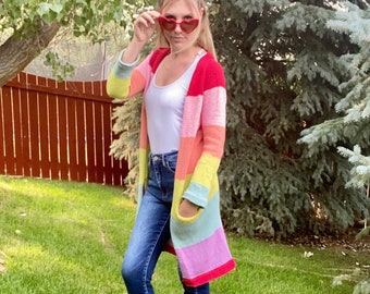 Puede incluir: Una mujer lleva un cárdigan a rayas arcoíris con un top blanco sin mangas, pantalones vaqueros azules y sandalias marrones. Lleva unas gafas de sol en forma de corazón en la mano.
