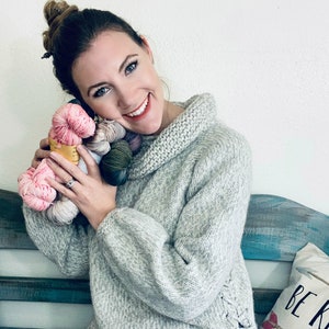 May include: A woman wearing a gray knitted sweater with a cowl neck and a side lace detail. She is holding a bundle of colorful yarn in her hands. The yarn is in shades of pink, green, and gray.