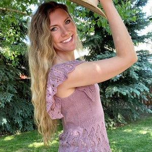 May include: A woman wearing a mauve knitted top with a delicate lace pattern and a wide-brimmed straw hat. She is standing in a grassy area with trees in the background.
