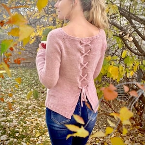 May include: A pink knitted sweater with a lace-up back detail. The sweater is worn over blue jeans and the model is standing in a field of fallen leaves.