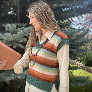 May include: A striped knit vest in shades of orange, teal, and cream. The vest is worn over a cream-colored button-down shirt. The vest has a V-neck and a green border at the bottom and around the armholes. A beaded necklace is worn.