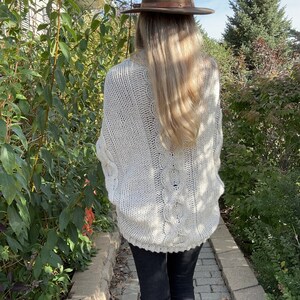 May include: A woman wearing a white cable knit sweater with a brown fedora hat and black pants.