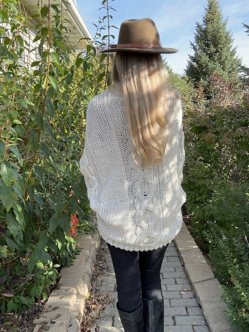 May include: A woman wearing a white cable knit sweater with a brown fedora hat and black pants.