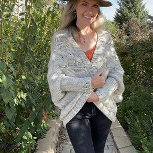 May include: A woman wearing a light gray cable knit cardigan with a brown fedora hat. She is wearing black jeans and a red tank top.