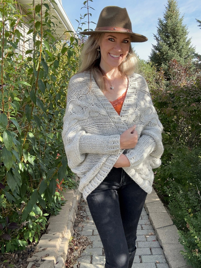 May include: A woman wearing a light gray cable knit cardigan with a brown fedora hat. She is wearing black jeans and a red tank top.