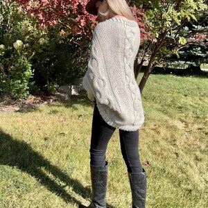 May include: A woman wearing a light gray cable knit sweater with a brown fedora hat and black jeans and boots.