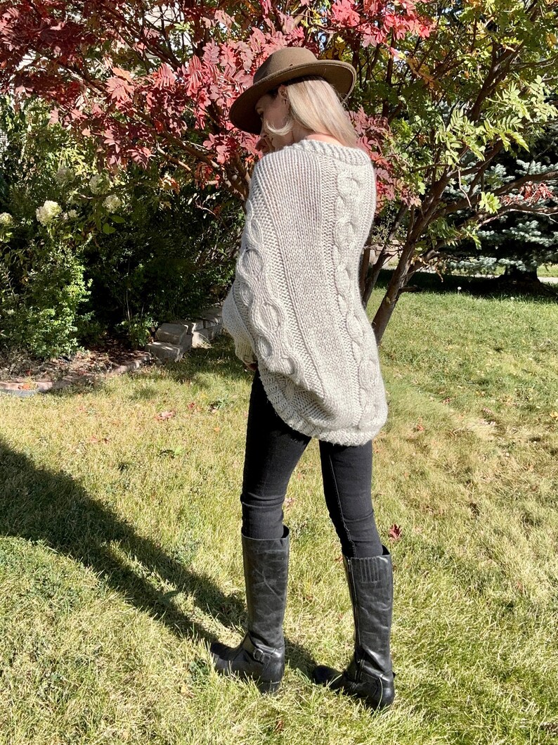 May include: A woman wearing a light gray cable knit sweater with a brown fedora hat and black jeans and boots.
