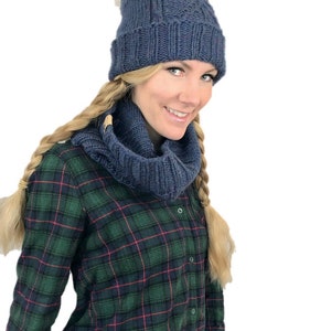 May include: A blue knitted hat with a white pom pom and a matching blue knitted scarf. The hat and scarf are worn by a person with long blonde hair and a plaid shirt.