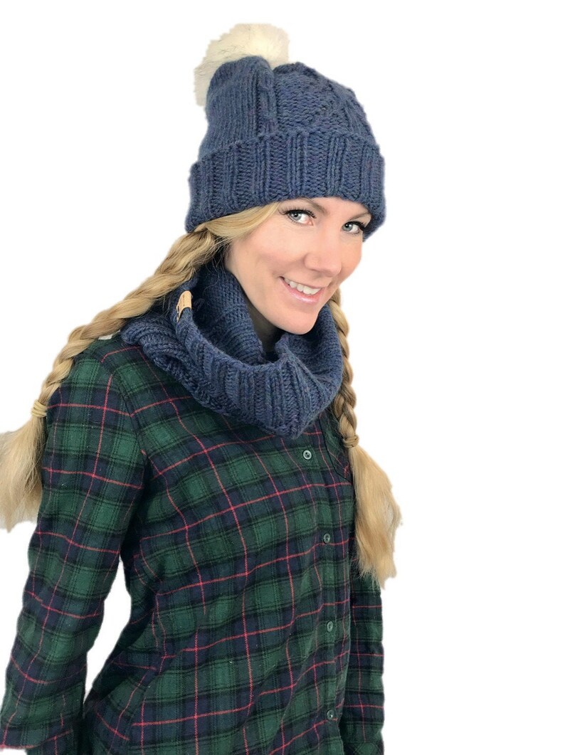 May include: A blue knitted hat with a white pom pom and a matching blue knitted scarf. The hat and scarf are worn by a person with long blonde hair and a plaid shirt.