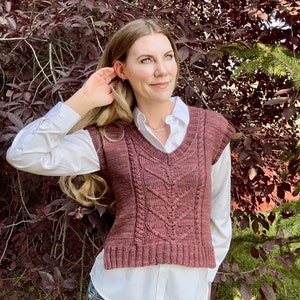 Könnte beinhalten: Eine maronfarbene, ärmellose Strickweste mit Kabelmuster und V-Ausschnitt. Die Weste wird über einem weißen Hemd mit Knöpfen und einer blauen Jeans getragen.