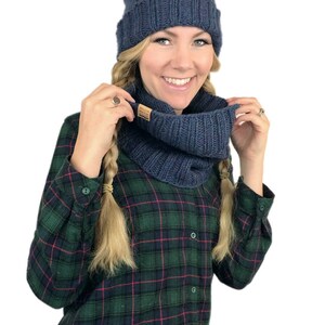May include: A blue knitted hat with a white pom pom and a matching blue knitted infinity scarf. The hat and scarf are worn by a person with blonde hair and a plaid shirt.