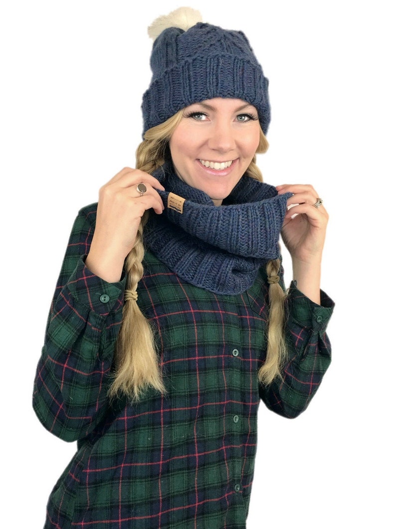 May include: A blue knitted hat with a white pom pom and a matching blue knitted infinity scarf. The hat and scarf are worn by a person with blonde hair and a plaid shirt.