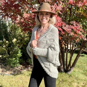 May include: A woman wearing a light gray cable knit cardigan with a brown fedora hat. She is standing in front of a tree with red leaves.