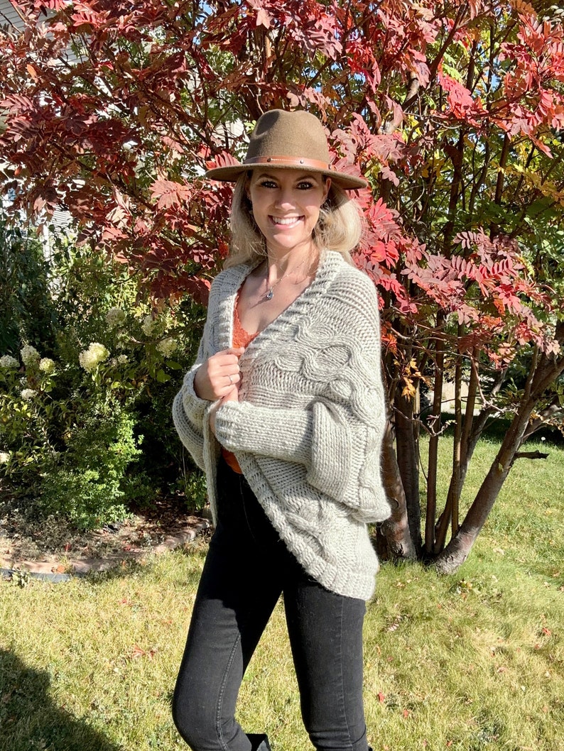 May include: A woman wearing a light gray cable knit cardigan with a brown fedora hat. She is standing in front of a tree with red leaves.