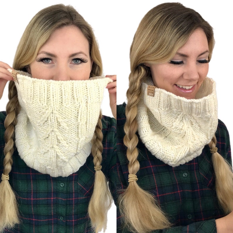 May include: A white knitted cowl neck scarf with a cable knit pattern. The scarf is worn by a person with long blonde hair in two braids.