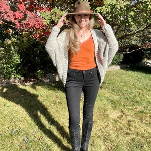 May include: A woman wearing a light gray knitted cardigan, a rust-colored tank top, black skinny jeans, and black knee-high boots. She is wearing a brown fedora hat and smiling.