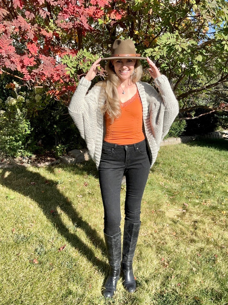 May include: A woman wearing a light gray knitted cardigan, a rust-colored tank top, black skinny jeans, and black knee-high boots. She is wearing a brown fedora hat and smiling.