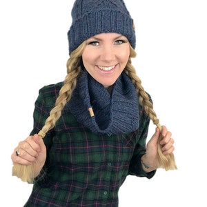 May include: A woman wearing a blue knit hat with a white pom pom and a matching blue knit scarf. She is wearing a green and black plaid shirt and has her hair in two braids.